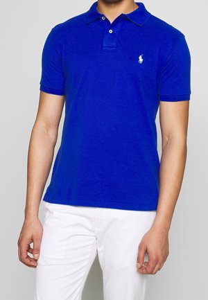 Man wearing a bright blue short-sleeve polo shirt with white embroidered logo on left chest and white pants against a plain background.