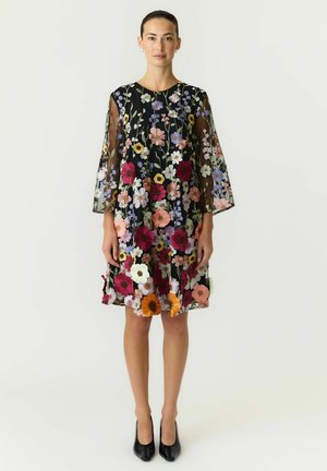 Femme portant une robe noire transparente longueur genou avec une broderie florale multicolore et des manches trois-quarts, associée à des talons noirs.
