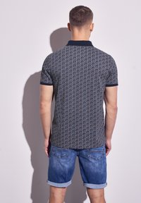 Camisa polo en color marino con un patrón geométrico, mangas cortas y un cuello en contraste. Combinada con shorts de denim enrollados. Vista trasera.