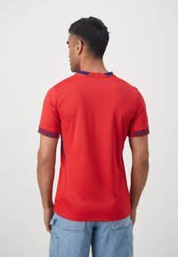 Maglia sportiva rossa con tessuto texturizzato, dettagli blu sul colletto e sui bordi delle maniche, e design aderente. Nessun logo o motivo visibile.