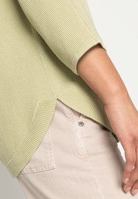 Pull vert clair en tricot avec un motif texturé et des manches trois-quarts, associé à un pantalon beige avec un détail de bouton visible.