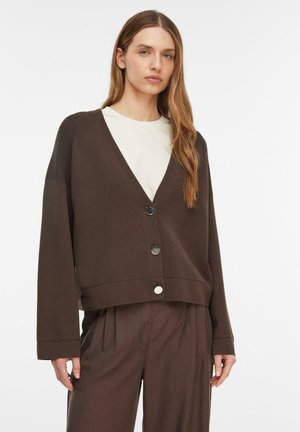 Kobieta ubrana w luźny, ciemnobrązowy rozpinany kardigan na białej koszuli, do tego brązowe plisowane spodnie, stojąca na jednolitym białym tle.