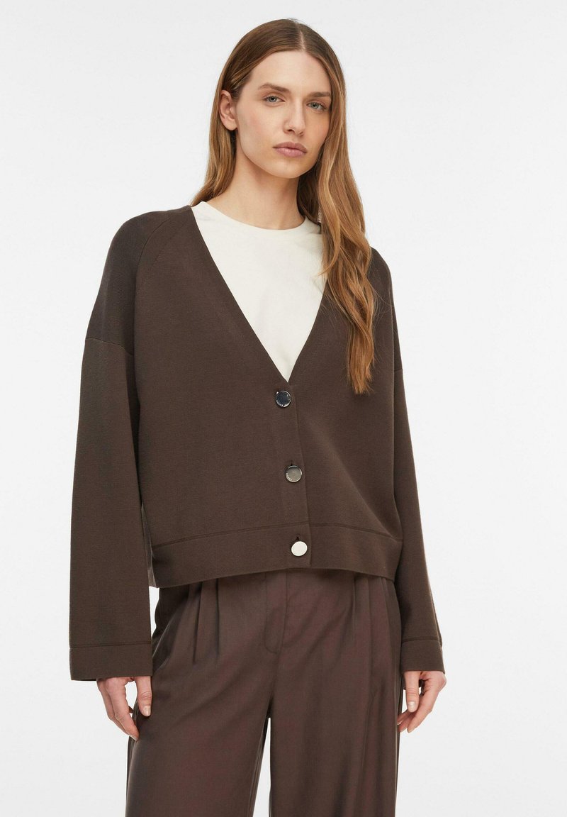 Frau trägt eine locker sitzende, dunkelbraune, geknöpfte Strickjacke über einem weißen Hemd mit passenden braunen, plissierten Hosen vor einem schlichten weißen Hintergrund.