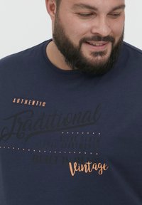 Marineblå T-skjorte i bomullsblanding med trykt tekst i svart og kobber. Teksten inkluderer "Autentisk", "Tradisjonell" og "Vintage."