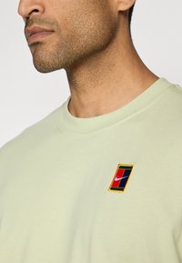 Light green short-sleeve T-shirt with a round neckline and embroidered rectangular logo in red, blue, and yellow on the left chest.