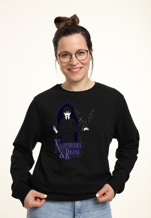 Donna sorridente con occhiali e capelli raccolti in un bun indossa un felpa nera con un personaggio gotico che presenta un corvo e la scritta "Nightshades & Ravens".