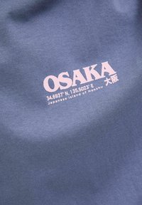 Mėlynas medvilninis audinys su žodžiu "OSAKA" rožin�ės spalvos, koordinatėmis ir tekstu "Japonijos sala Honšū" apačioje. Paprasta, modernaus dizaino tipografija.