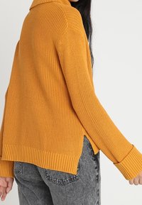 Femme portant un pull en maille jaune moutarde avec des fentes latérales et des manches longues, associé à un jean gris délavé, sur un fond uni.