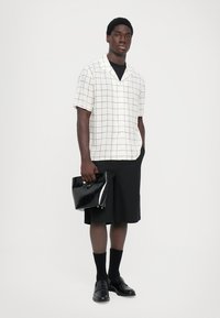 Witte shirt met korte mouwen en zwart rasterpatroon, gecombineerd met een zwarte rok tot de knie, zwarte schoenen en een glanzende zwarte clutch.