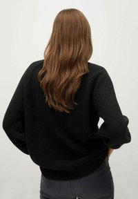 Osoba s dlouhými hnědými vlasy má na sobě volný černý svetr a tmavé kalhoty, stojí s rukama v bok, čelem k světle šedému pozadí.