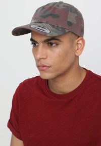 Cappellino mimetico in tonalità di marrone e verde, realizzato in tessuto con visiera piatta. Indossato con una maglietta a maniche corte in cotone rosso uniforme.