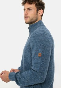 Blauer Strickpullover mit hohem Kragen, gerippten Bündchen und einem Leder-Logo-Patch am linken Ärmel. Er verfügt über ein strukturiertes, gesprenkeltes Muster.