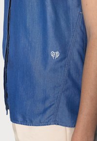 Modrá denimová košile s krátkým rukávem, s malým bílým výšivkovým logem srdce na levé straně blízko lemu. Hladká textura.