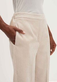 Donkerhuidige persoon in lichtbeige ribgebreide broek met elastische tailleband, hand in de zijzak, en witte top die erin gestoken is.