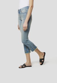 Lichtblauwe denim capri jeans met geborduurde details in blauwe en gouden patronen aan de zoom, met rafelige randen. Draagt deze met zwarte slides sandalen.