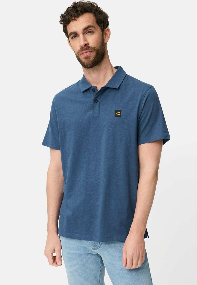Man met krullend haar en baard, gekleed in een blauw poloshirt met korte mouwen en lichtblauwe spijkerbroek, staand tegen een effen achtergrond.