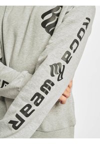 Grå bomulds sweatshirt med sort trykt tekst ned ad ærmerne og et diskret logo på fronten, med ribbede håndled og kant.