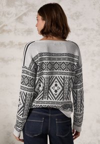 Pull gris avec des motifs géométriques noirs, col bateau et poignets côtelés, associé à un jean en denim foncé. Accent sur la texture et les détails de conception.