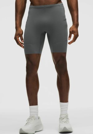 Mann trägt enganliegende dunkelgraue Sportshorts, weiße Socken und weiße Laufschuhe, steht vor einem einfarbigen hellen Hintergrund.