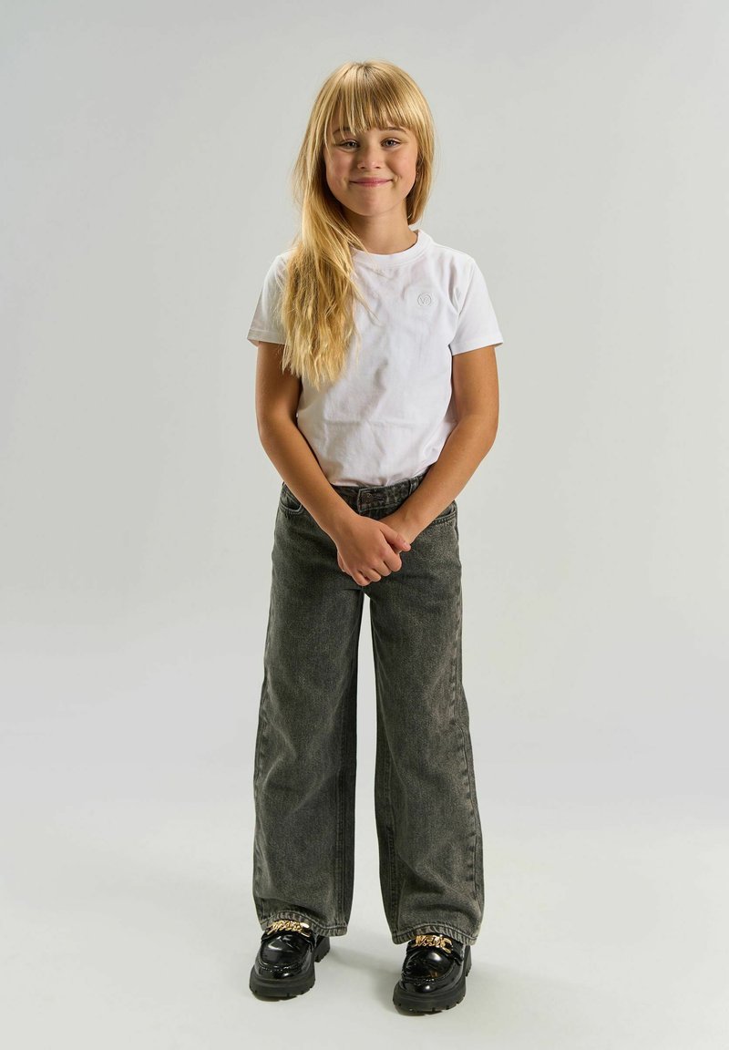 Jeune fille aux longs cheveux blonds, debout en souriant, portant un t-shirt blanc, un jean ample sombre et des chaussures noires avec des détails en chaîne dorée.