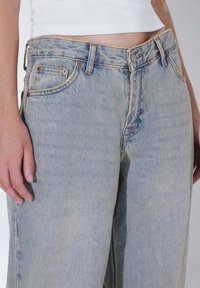 Ljusblå denimjeans med avslappnad passform, som har kopparbeslag, framfickor och synliga sömnadsdetaljer längs midjan.