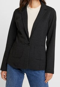 Svart skräddarsydd blazer med vitt prickmönster, enkel knäppning, slagkragar och framfickor, kombinerad med en beige topp.