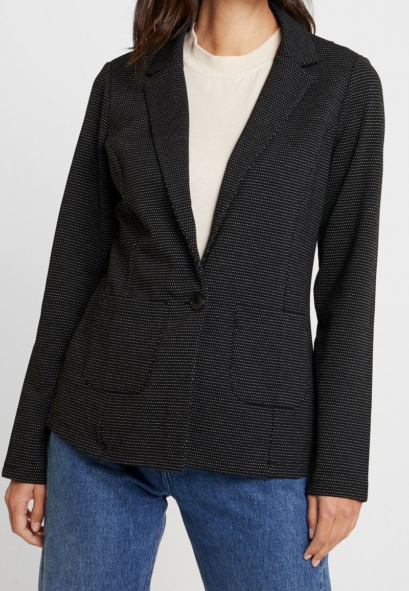 Svart skräddarsydd blazer med vitt prickmönster, enkel knäppning, slagkragar och framfickor, kombinerad med en beige topp.