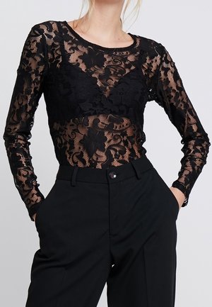 Kvinde iført en gennemsigtig sort blomstret blonder langærmet top over en sort bh, kombineret med højtaljede sorte bukser, hænderne i lommerne.