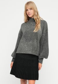 Pelēks, teksturēts džemperis ar augstu uzmavu un vaļīgām piedurknēm, apvienots ar melnu plīša svārku ar kontrastējošiem šuvumiem.