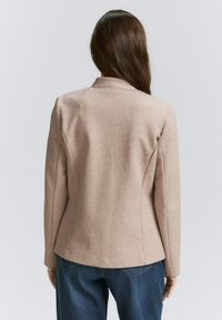 Blazer beige su misura con una silhouette aderente, rever a punta e cuciture sulle spalle. Tessuto testurizzato, maniche lunghe e dettagli di cucitura sottili.