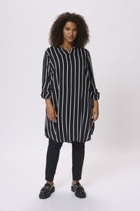 Donna con capelli ricci che indossa una tunica a righe verticali bianche e nere, pantaloni neri e mocassini neri, in piedi davanti a uno sfondo semplice.