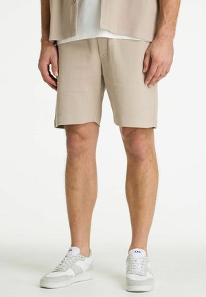 Moški v bež kratkih hlačah z teksturo, z belimi in bež supergami, stoji z rokami sproščenimi ob telesu pred belim ozadjem.