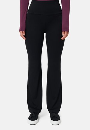 Person trägt schwarze Schlaghosen, schwarze Sneakers mit weißen Sohlen und ein langärmeliges lila Oberteil, steht vor einem einfarbigen Hintergrund.