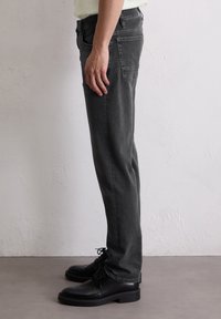 Zijaanzicht van een persoon in vervaagde zwarte jeans en zwarte veterschoenen die tegen een witte textuur muur staat.