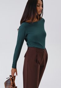 Vrouw in een teal lange mouw top en bruine hoge taillebroek, die een geruite handtas met leren hengsels vasthoudt tegen een effen achtergrond.