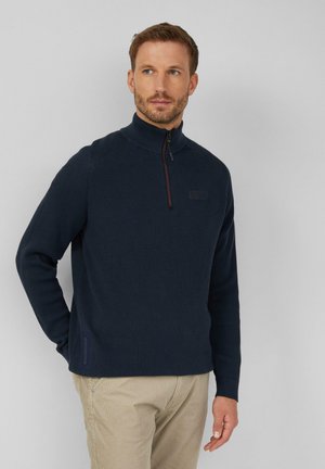 Pull en tricot bleu marine avec un col montant, ouverture demi-zippée, accents latéraux et tissu texturé. Porté avec un pantalon beige.