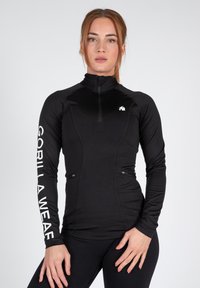 Zwarte lange mouwen sporttop met hoge kraag, ritssluiting en logo op de borst en mouw. Heeft een aansluitend ontwerp en twee ritssluitingen zakken.