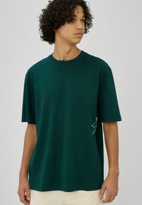 Jeune homme aux cheveux bouclés portant un T-shirt vert foncé surdimensionné avec un petit motif blanc sur le côté, pantalon beige.