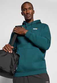 Felpa con cappuccio teal, dotata di tasca a marsupio, polsini a costine e logo Puma ricamato. Il modello tiene in mano una borsa da ginnastica nera.