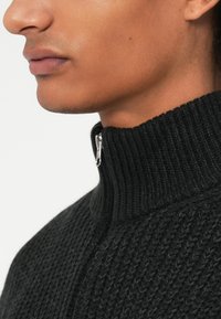 Maglione nero a maglia con colletto alto e zip frontale. La trama è a coste con un motivo sottile, che enfatizza calore e comfort.