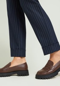 Mocassini in pelle marrone con finitura testurizzata e suola nera rigata, abbinati a pantaloni a righe blu navy. I pantaloni hanno un orlo risvoltato.