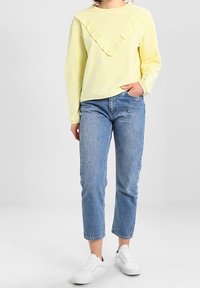 Felpa gialla con design a V arricciato, abbinata a jeans cropped blu chiaro e sneakers bianche. Tessuto dalla superficie liscia.