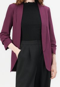 Lila skräddarsydd blazer med öppen front, trekvartsärmar, slätt tyg, bärs över en svart ribbad topp och högmidjade svarta byxor.