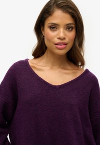 Fioletowy sweter w serek wykonany z miękkiej, teksturowanej tkaniny; charakteryzuje się luźnym krojem, szerokimi rękawami i lekko zniszczonym dekoltem.