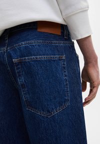 Jeans de mezclilla de color azul oscuro con un acabado texturizado, que cuentan con un parche de cuero marrón en la cinturilla y dos bolsillos traseros con detalles de costura.