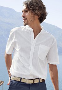 Weißes Kurzarmhemd mit Stehkragen, mit einer Brusttasche. Getragen mit einem gewebten beigen Gürtel und blauen Hosen. Malerischer Hintergrund im Freien.