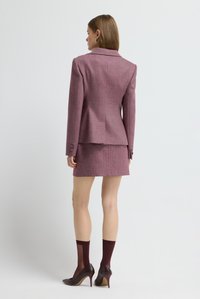 Woman wearing a mauve herringbone blazer and matching skirt, paired with dark burgundy sheer socks and high heels, standing against a plain background.