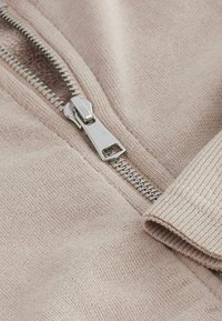 Světle růžová pletená látka svetru se stříbrným kovovým zipem a žebrovanými manžetami. Detailní záběr zaměřený na zip a texturu.