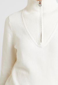 Pull à col roulé crème en tricot avec un col en V profond et une texture côtelée, doté d'un demi-zip au niveau du col.
