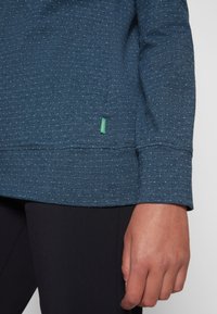 Gros plan sur la main d’une personne reposant à ses côtés, portant un haut bleu à manches longues tacheté et un pantalon noir.
